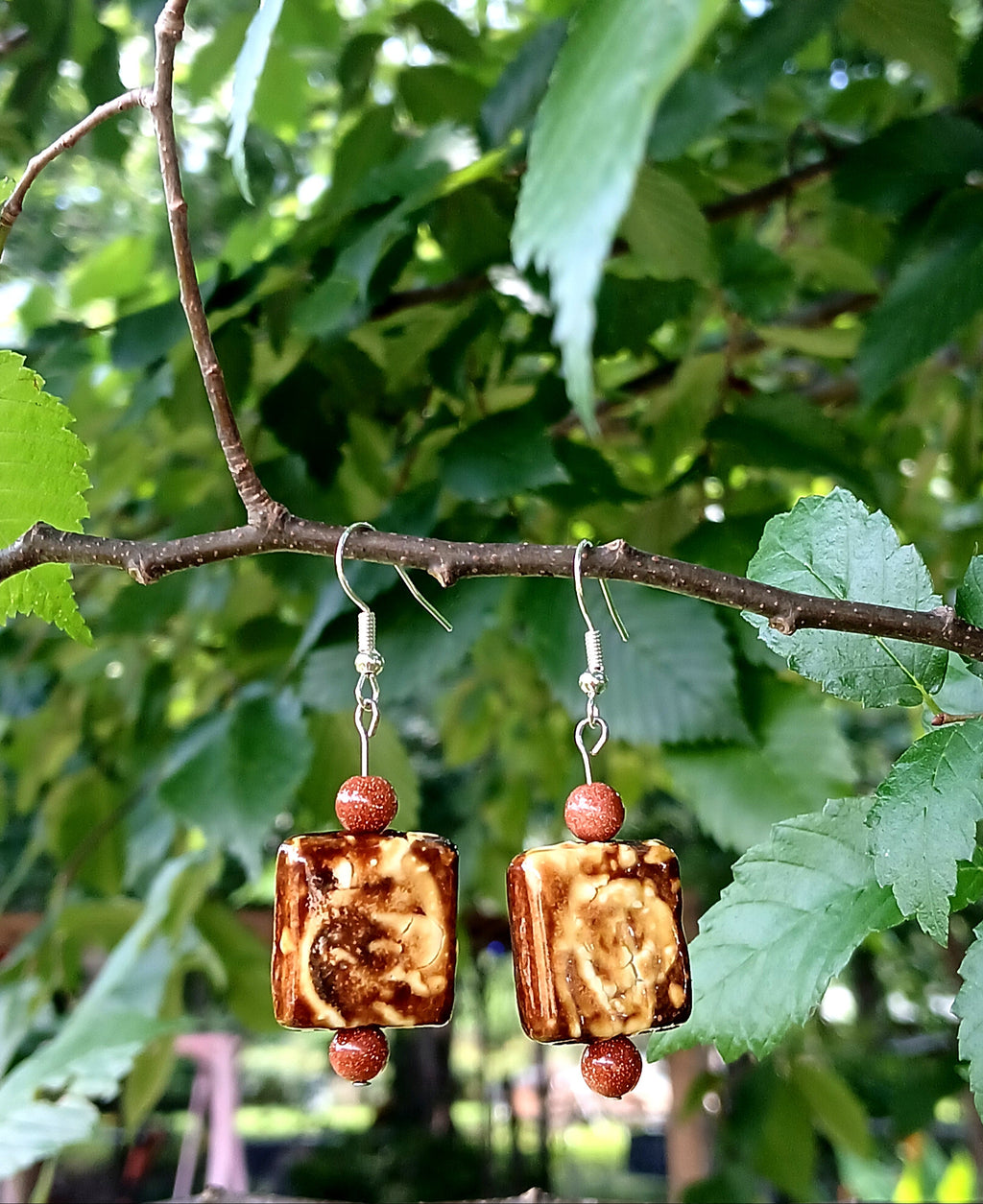 Pair of earrings with brown square beads and red beads hanging on a branch with green leaves.