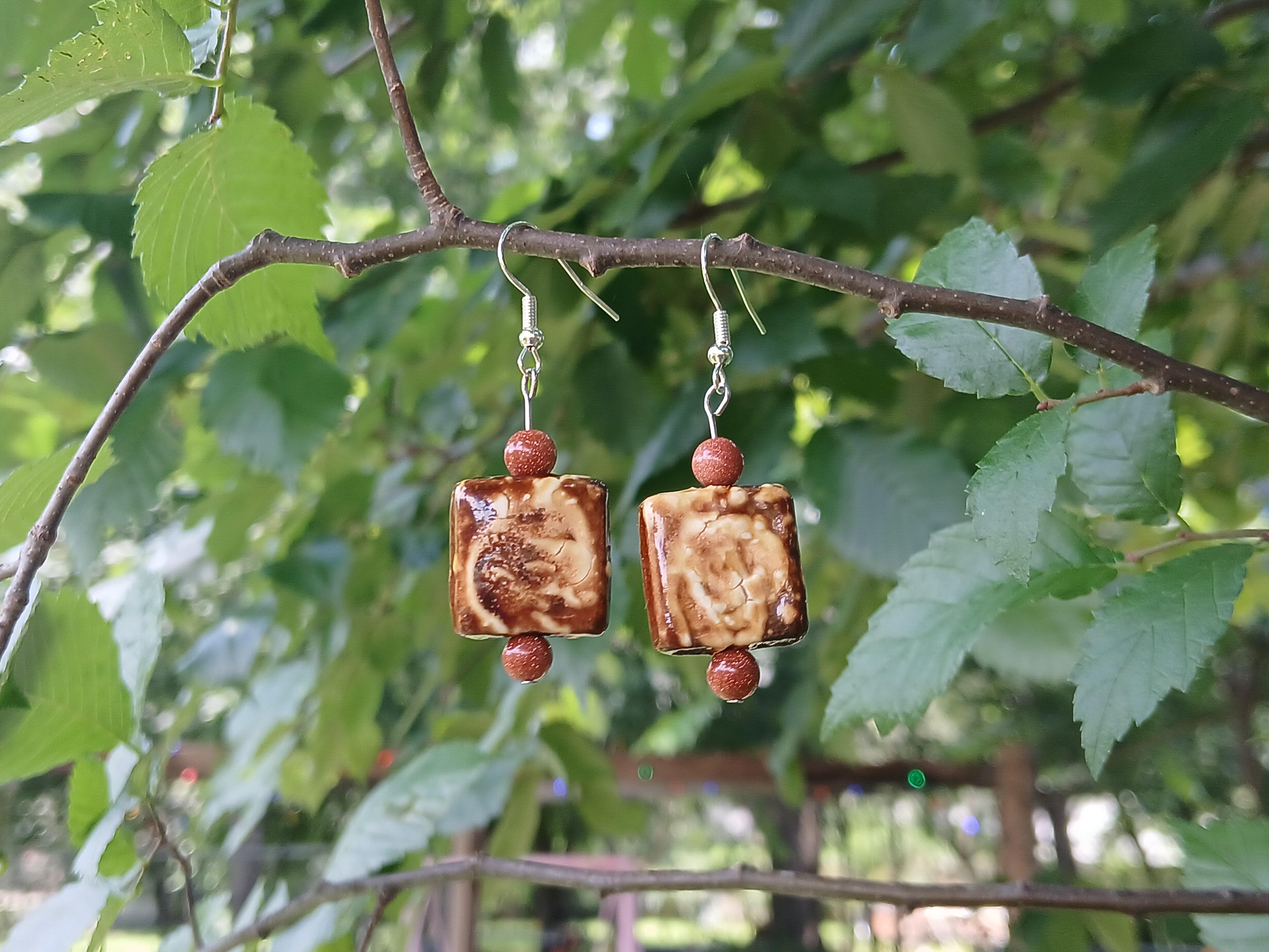 Pair of square-shaped earrings with red beads hanging on a branch with green leaves.