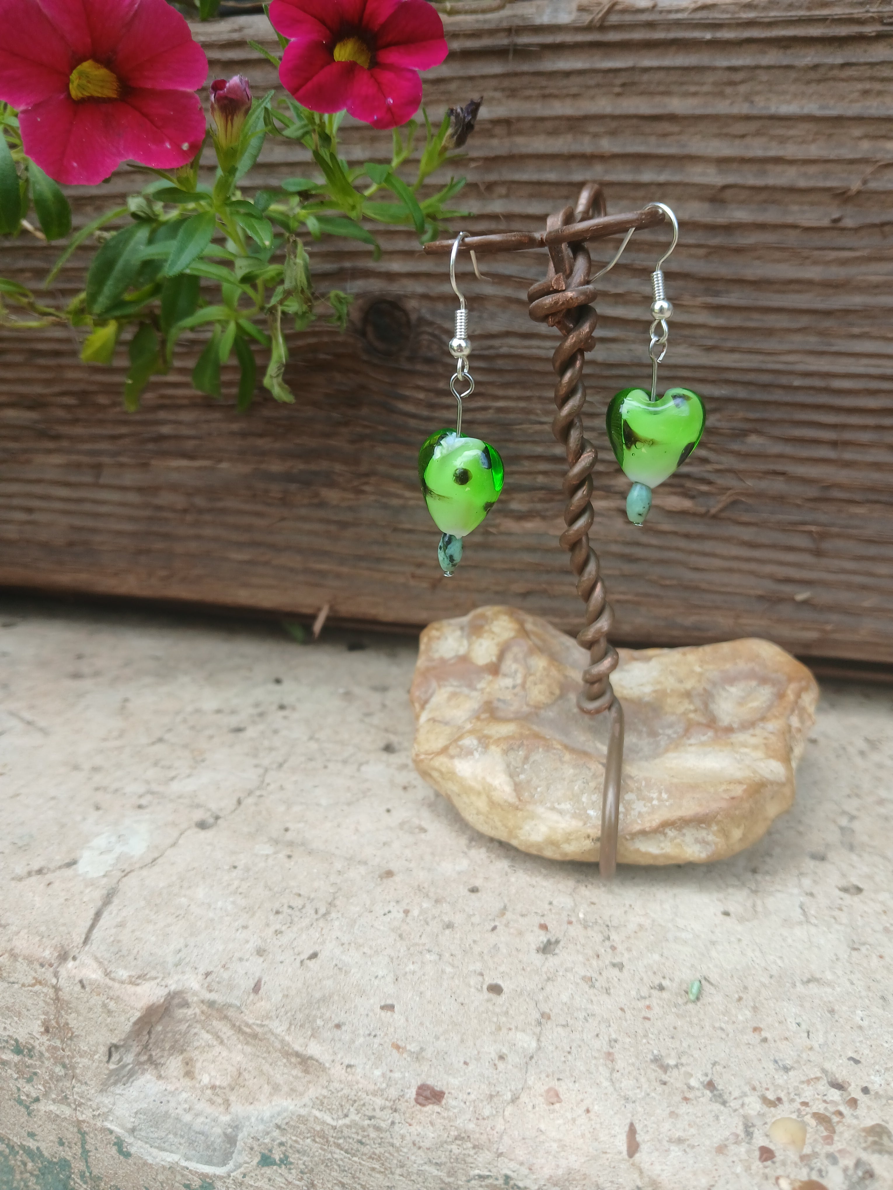 Green heart-shaped earrings on a stone stand with pink flowers in the background
