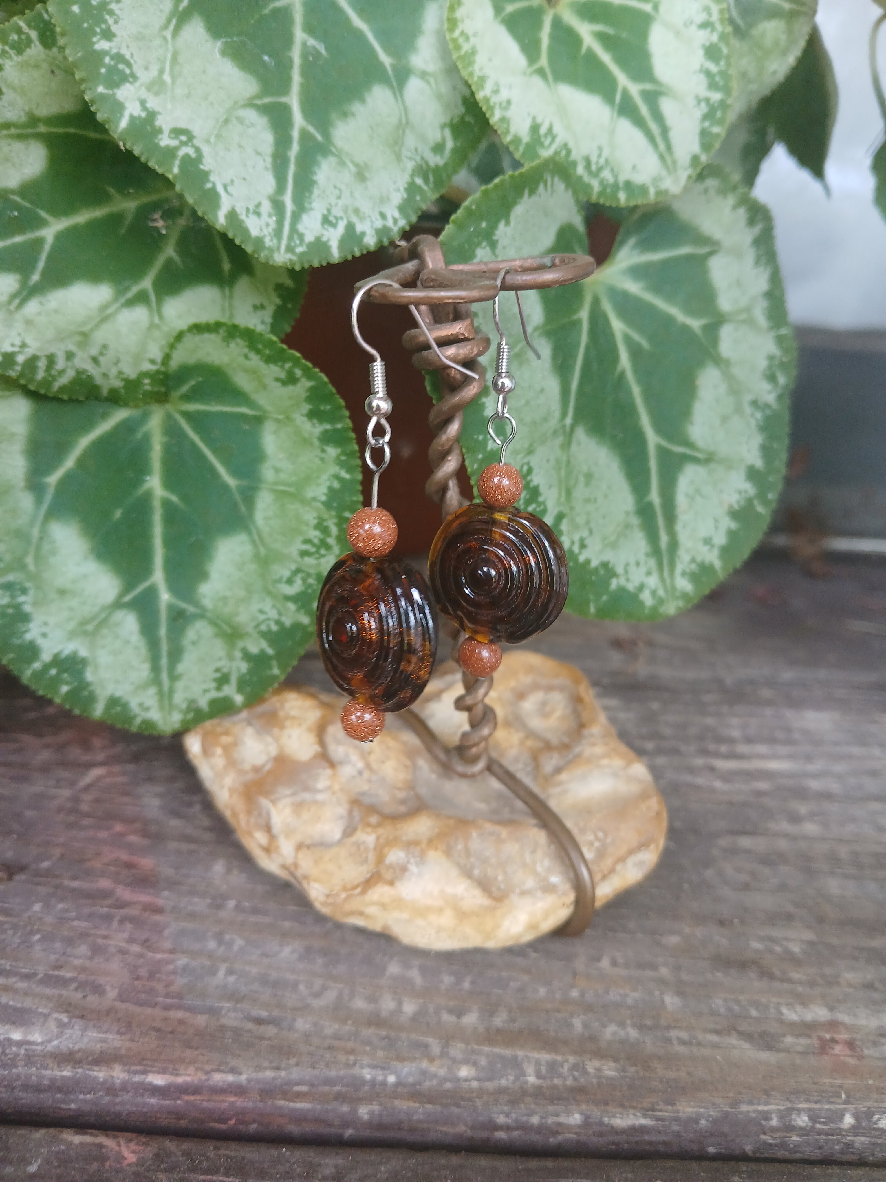 Pair of brown beaded earrings on a stone surface with green leaves in the background