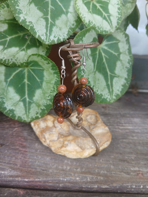 Pair of brown beaded earrings on a stone surface with green leaves in the background