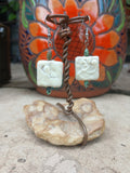 Pair of earrings with white square stones and brown wire on a textured surface.