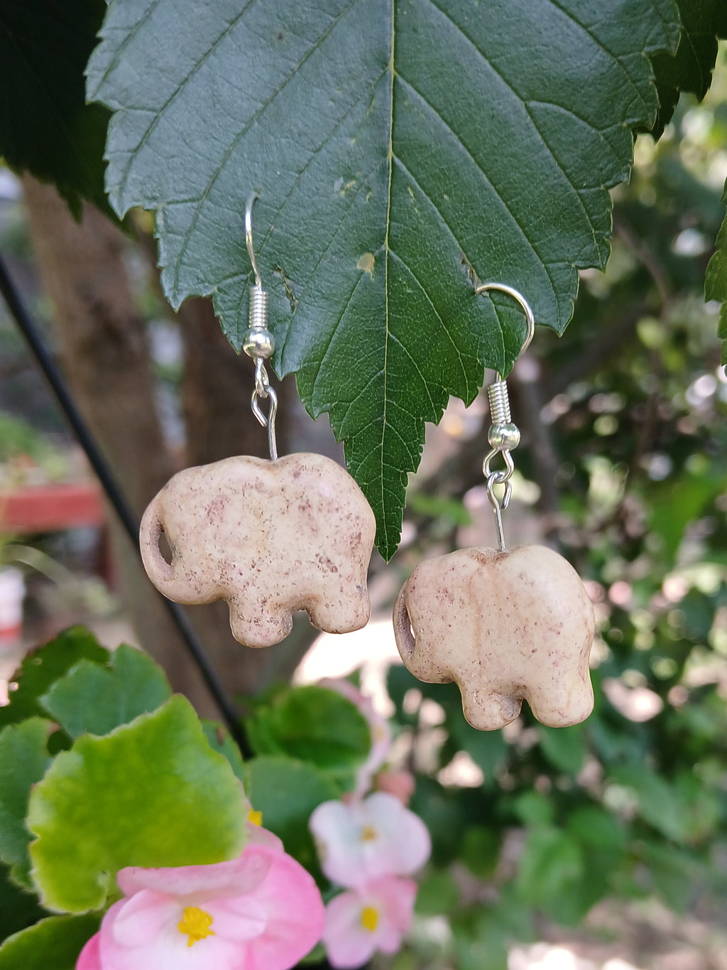 Two beige stone-like earrings hanging on a green leaf with blurred garden background