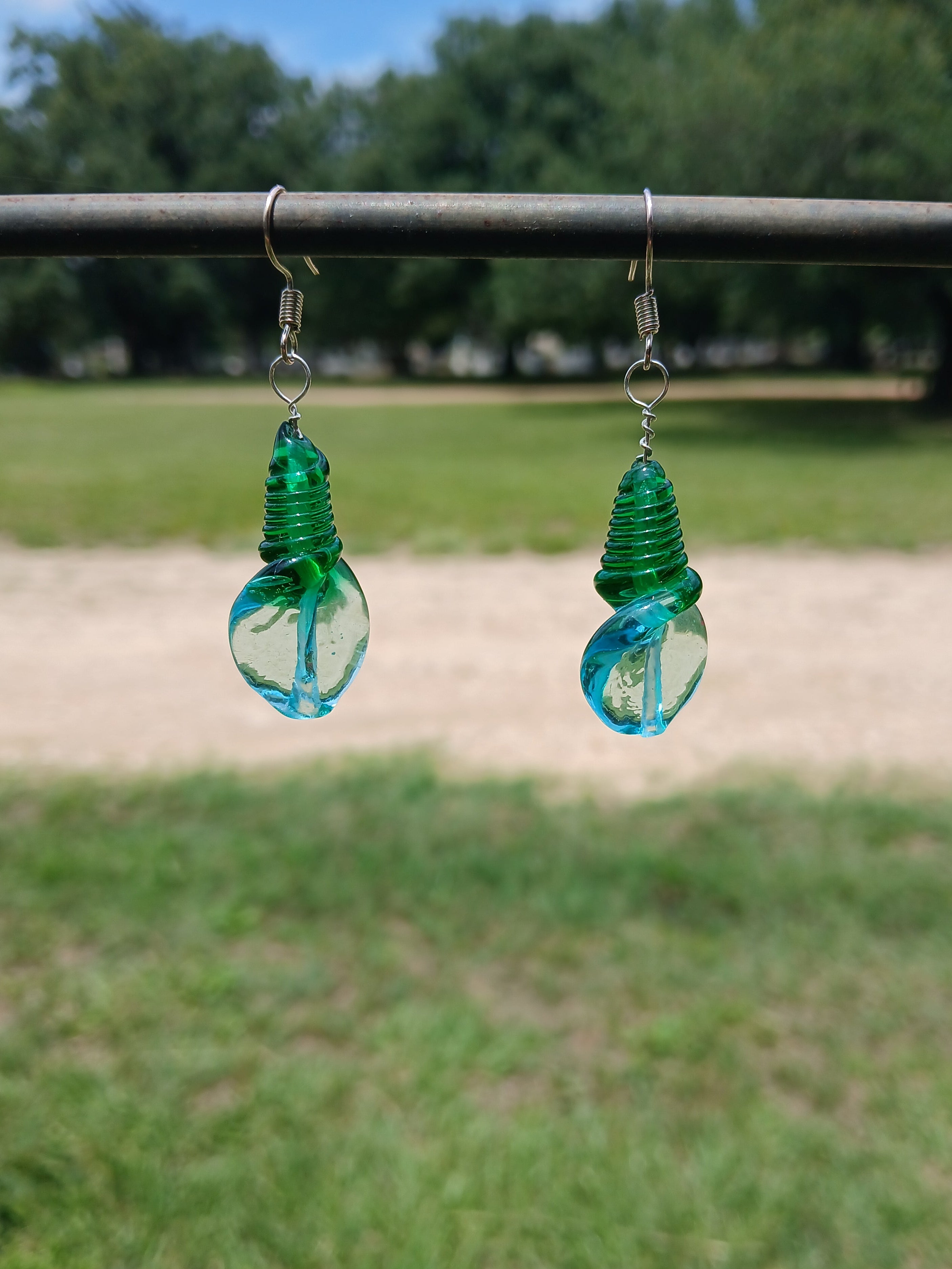 Green glass earrings hanging from a wire with a blurred road and trees in the background