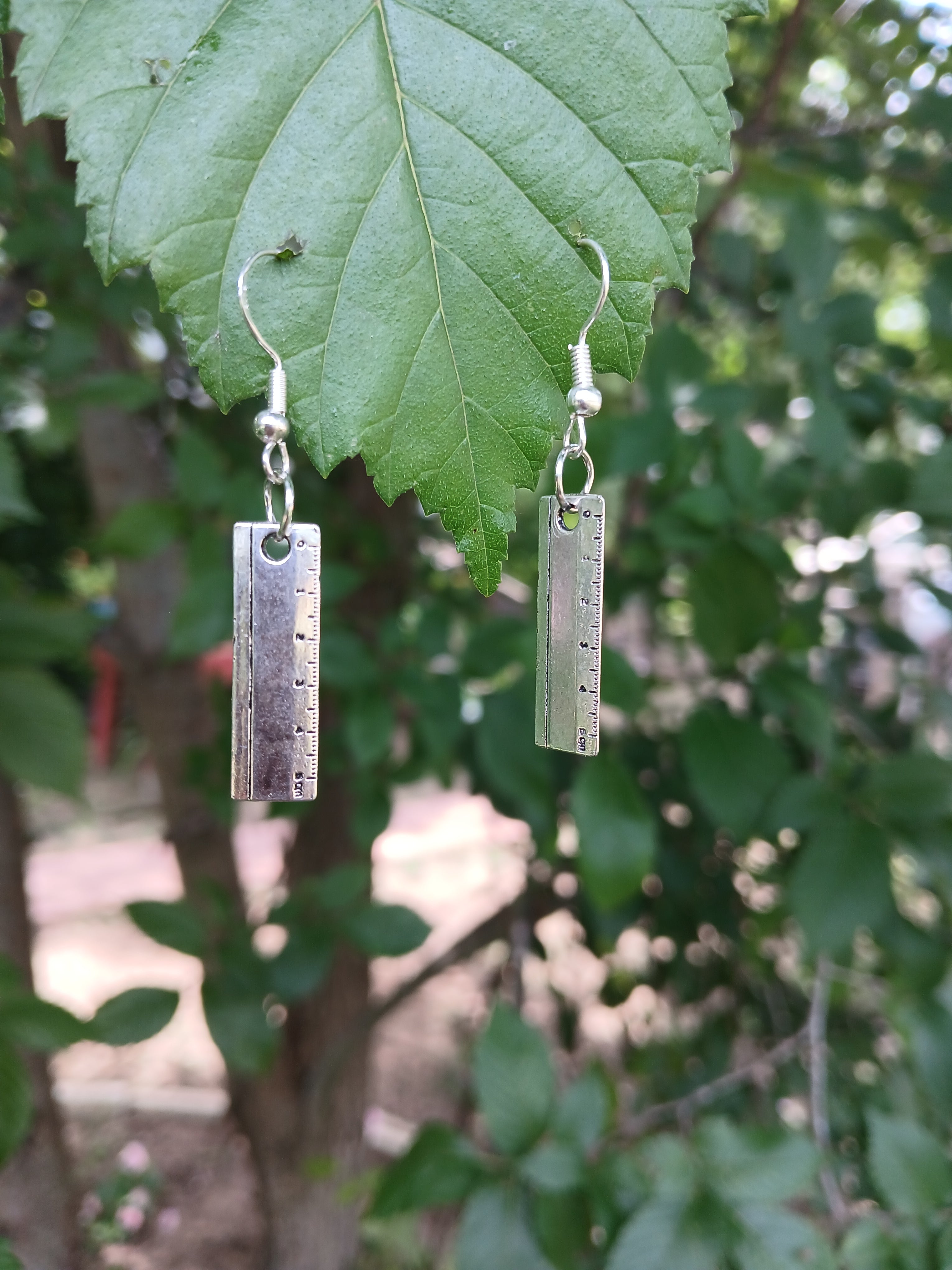 Silver ruler-shaped earrings on a green leaf with a blurred natural background